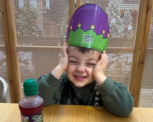 Child wearing a colorful hat with 'Fruit Shoot' branding, sitting at a table with a bottle of the product.